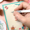 A hand holding a silver and white mechanical pencil writes on a cream-coloured notepad with a teal border and cute sewing-themed illustrations. The background is a soft pink surface with sewing supplies like spools of thread, scissors, and fabric patches.