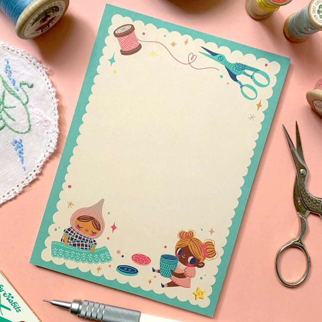 A square notepad with a scalloped teal border and whimsical sewing-themed illustrations, styled on a peach background with thread spools, scissors, a pen, and other crafting props.