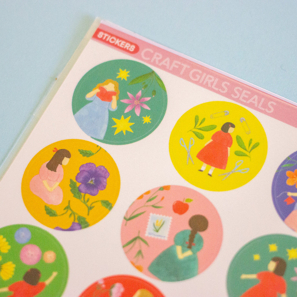 A close-up of a sticker sheet titled 'STICKERS CRAFT GIRLS SEALS' showing several colourful circular stickers with illustrated girls and flowers on a pale blue background.