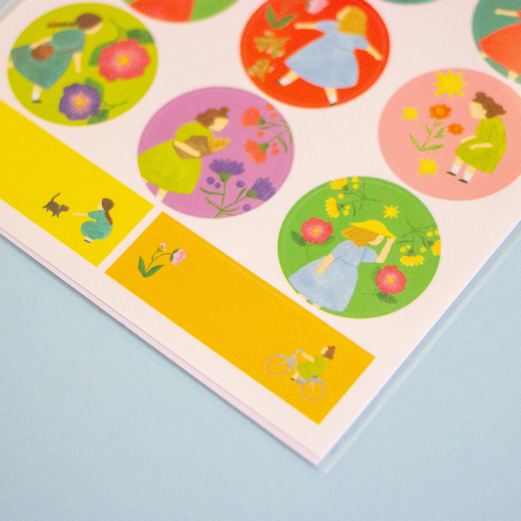 A close-up of a printed sticker sheet or art print featuring colourful circular and rectangular illustrations of girls in dresses with flowers, bicycles, and small animals, placed on a light blue surface.