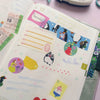 A close-up of a decorated planner or journal page with colourful washi tape strips, sticky notes, and penguin illustrations, photographed on a soft lilac surface.