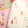 A weekly planner spread in a clear plastic sleeve, decorated with cute pastel illustrations of girls, flowers, and hearts, shown alongside sticker sheets and a pen on a pink background.
