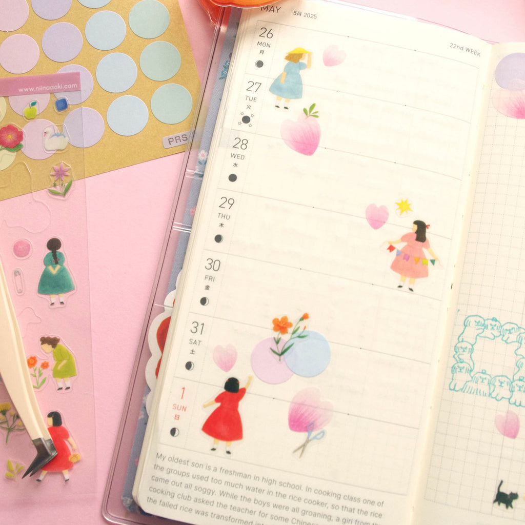 A weekly planner spread in a clear plastic sleeve, decorated with cute pastel illustrations of girls, flowers, and hearts, shown alongside sticker sheets and a pen on a pink background.