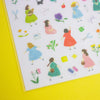 A sheet of colourful sticker illustrations featuring girls in dresses, flowers, and small accessories, photographed on a bright yellow background.