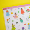 A sheet of craft stickers featuring cute cartoon girls in colourful dresses and various small decorative items, displayed on a bright yellow background.