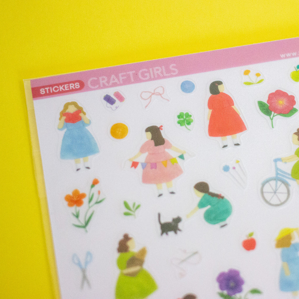 A sheet of craft stickers featuring cute cartoon girls in colourful dresses and various small decorative items, displayed on a bright yellow background.