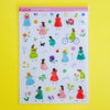 A sheet of craft stickers featuring cute cartoon girls in colourful dresses and various small decorative items, displayed on a bright yellow background.