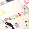 A close-up of a decorative planner or journaling strip with illustrated penguins, flowers, small figures, and pastel motifs on a white grid paper background.