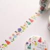 A roll of decorative washi tape with a white background and colourful illustrations of girls in dresses and flowers, shown partially unrolled on a light surface. A round label reading 'CRAFT GIRLS' is visible near the bottom right.