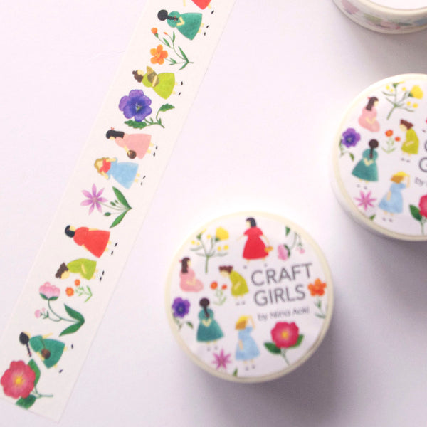A close-up of decorative paper strips featuring a repeating illustration of girls in colourful dresses walking beside flowers, photographed on a white surface.