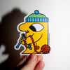 A hand holds a die-cut sticker featuring a yellow cartoon character wearing a green and blue beanie and holding a red roll of yarn, against a plain white wall with a cast shadow.