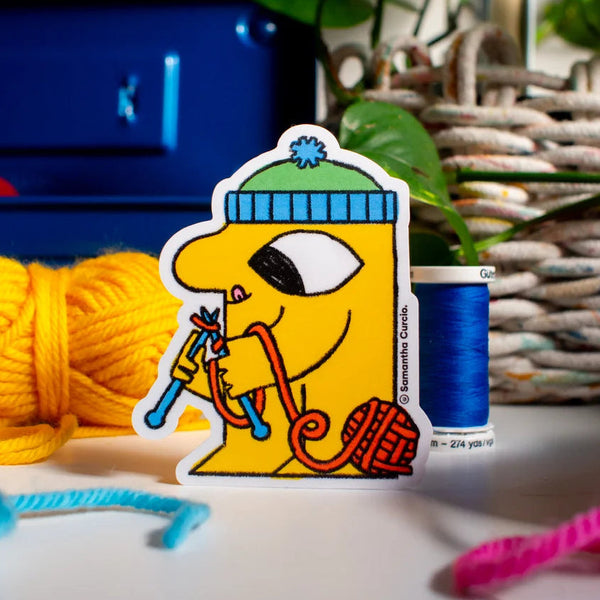 A die-cut sticker featuring a yellow cartoon character knitting with blue and red yarn, styled on a tabletop with knitting supplies including a skein of yellow yarn, blue thread, and a plant in the background.