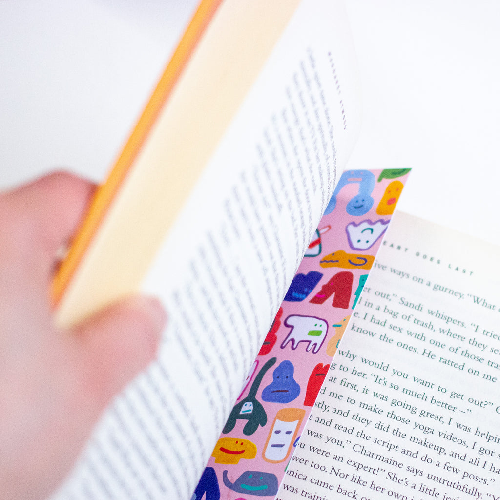 A close-up of a hand holding an open book, with a colourful bookmark featuring cartoon animal faces printed on it.