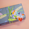 A closeup of the creatures gift tag. This print features a fun and whimsical gathering of quirky, wild characters. The gift tag is attached to a green string on a gift box.