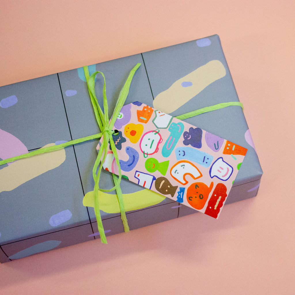 A closeup of the creatures gift tag. This print features a fun and whimsical gathering of quirky, wild characters. The gift tag is attached to a green string on a gift box.
