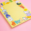A pastel yellow lined notepad with cute cartoon character illustrations around the border, photographed on a soft pink background. The brand name 'shuh.'