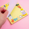 A hand holds a white sheet over a pastel yellow notepad decorated with colourful cartoon characters, set against a solid pink background.