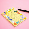 A yellow notepad with whimsical cartoon characters and handwritten text, styled on a pink background next to a black pen.