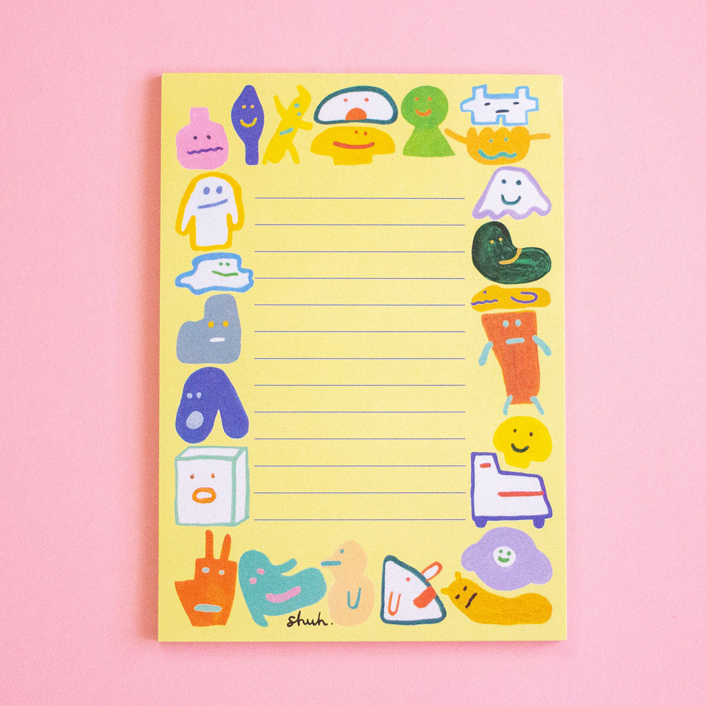 A yellow lined notepad with whimsical cartoon character illustrations around the border, photographed on a soft pink background. The brand name 'shuh.