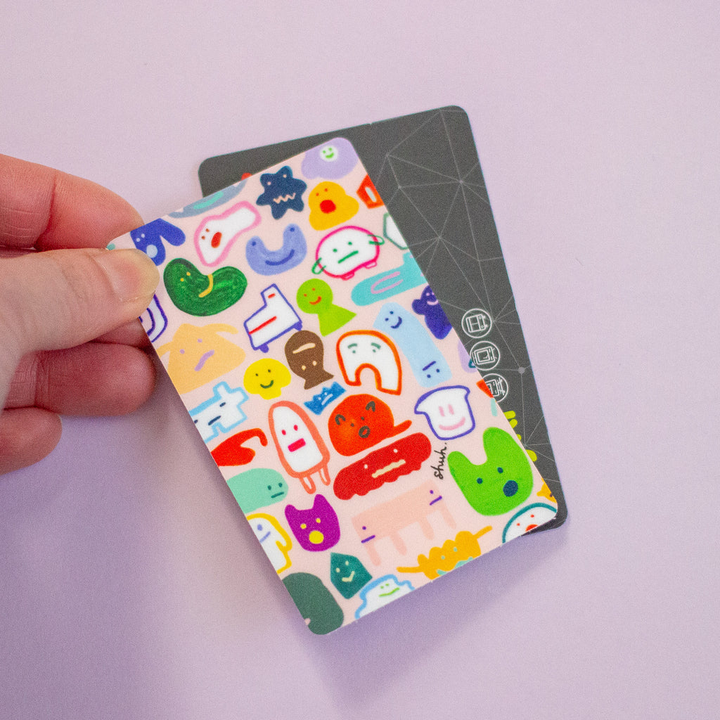 A hand holds a rectangular sticker with a colourful cartoon character print over a dark grey card on a light lavender background.