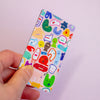 A hand holding a sticker with a colourful cartoon character pattern against a soft light purple background.