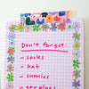 A close-up of a pink grid notepad decorated with a colourful floral border and a patterned washi tape strip at the top. Handwritten pink text on the pad reads, 'Don't forget: socks hat sunnies earplugs'.
