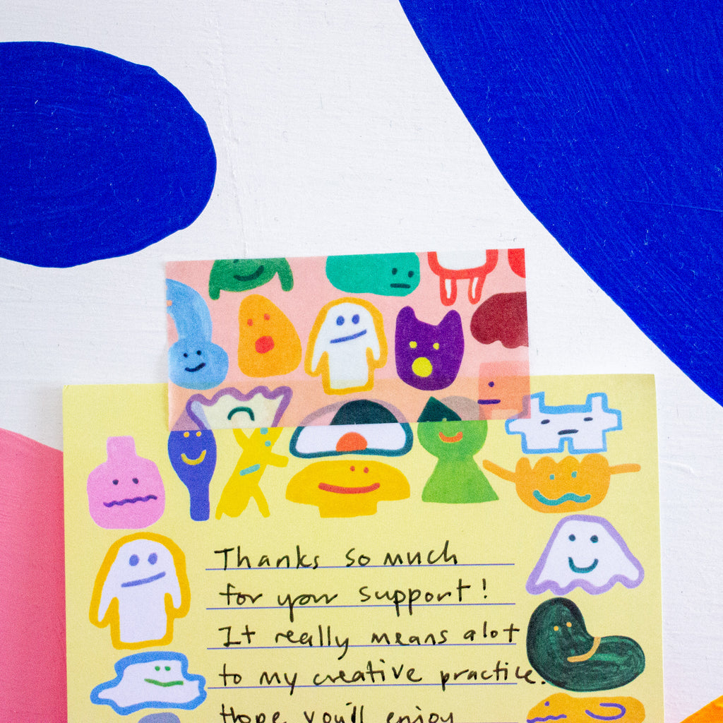 A close-up of a colourful tape with cartoon ghost-like illustrations and handwritten text on a pale yellow notepad, layered over a colourful wall. 