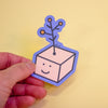 A hand holding a die-cut sticker featuring a cute cartoon character with a yellow flower on its head, set against a plain light yellow background.