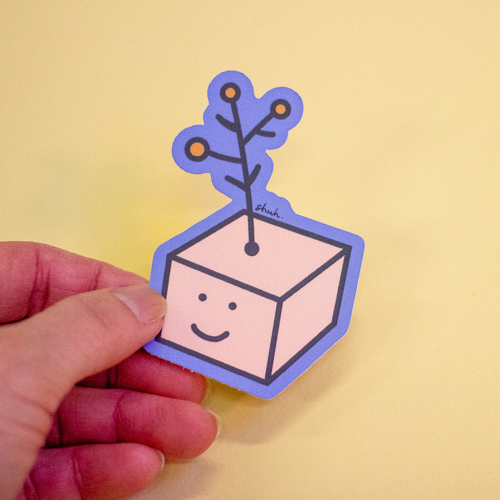 A hand holding a die-cut sticker featuring a cute cartoon character with a yellow flower on its head, set against a plain light yellow background.