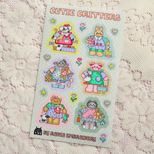 A sticker sheet titled 'CUTIE CRITTERS' featuring six cute pixel-art animal characters in pastel clothing, placed on a textured cream lace fabric background. 