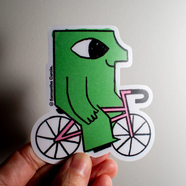 A hand holding a die-cut sticker featuring a green figure riding a pink bicycle against a plain light grey background.
