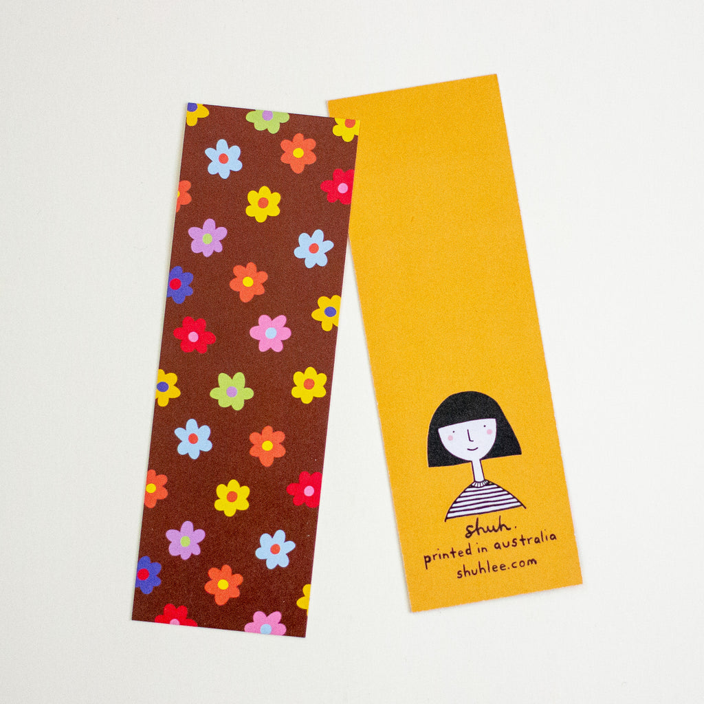 Two bookmarks on a white background. One bookmark has a colourful floral-patterned bookmark with pink, red, purple, orange, yellow, green, and blue flowers. The other one has a plain mustard background with shuh. logo and words on the bottom center. Both bookmarks are shown against a plain white background.