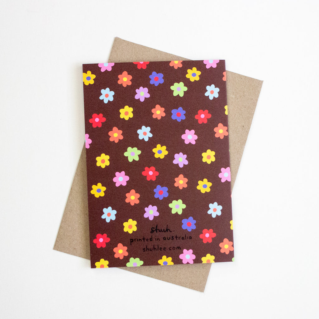 A card with colourful flower illustrations all over against a brown background with a shuh. logo on the bottom of the card on a kraft paper envelope. 