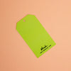 A bright lime green paper gift tag with a hanging hole, featuring the brand name 'shuh.' and website printed in black at the bottom, set against a solid peach background.