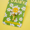 A smiling daisy enamel pin with a yellow face, white petals, and green leaves, displayed on its green floral backing card by Hannakin.