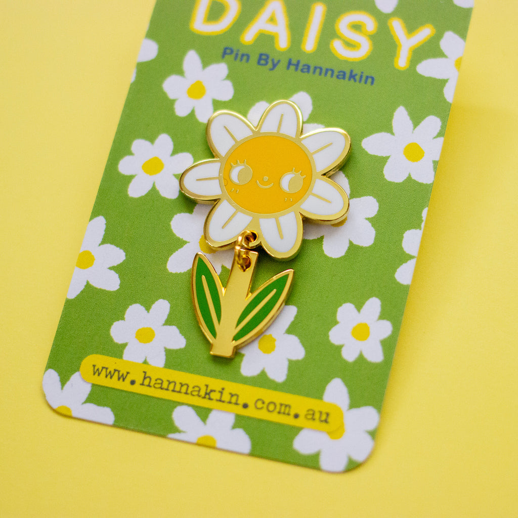 A smiling daisy enamel pin with a yellow face, white petals, and green leaves, displayed on its green floral backing card by Hannakin.
