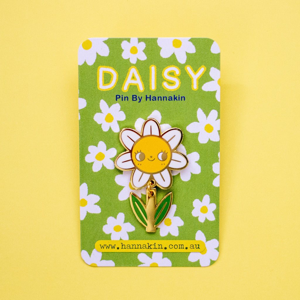 A daisy-shaped enamel pin with a smiling yellow face, white petals, and green leaves, mounted on a green floral backing card that reads 'DAISY Pin By Hannakin'.