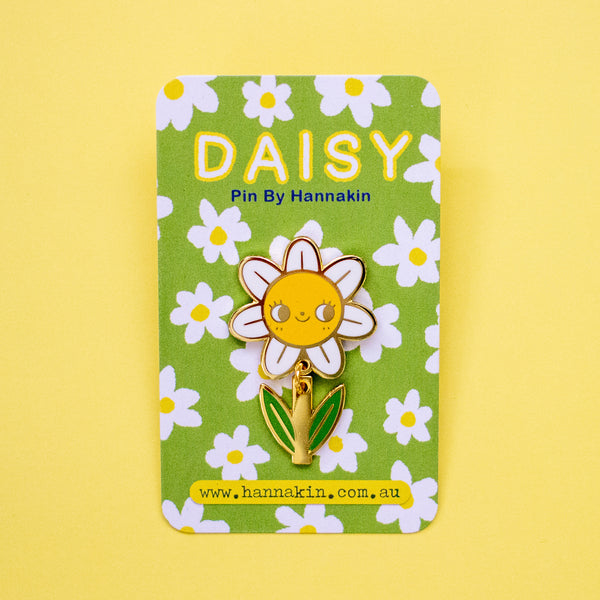A daisy-shaped enamel pin with a smiling yellow face, white petals, and green leaves, mounted on a green floral backing card that reads 'DAISY Pin By Hannakin'.