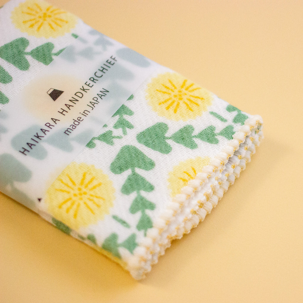 A folded white handkerchief with a green leaf and yellow flower pattern, featuring the brand name 'HAIKARA HANDKERCHIEF' printed on it, set against a solid pale yellow background.