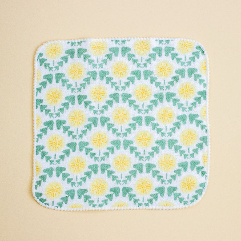 A square white fabric item with rounded corners, featuring a repeating pattern of yellow flowers and green leaves, finished with a scalloped yellow border, set against a solid beige background.
