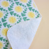 A close-up of a folded fabric item showing a white side with a green and yellow dandelion pattern on the front and a soft white fleece backing on the folded-over side.