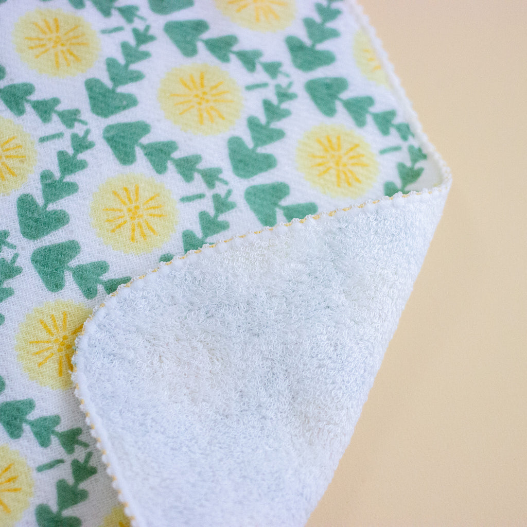 A close-up of a folded fabric item showing a white side with a green and yellow dandelion pattern on the front and a soft white fleece backing on the folded-over side.