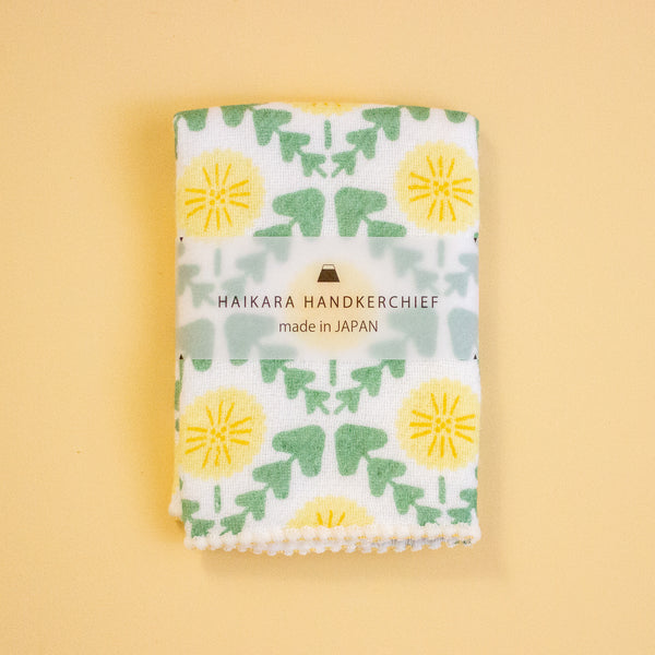 A folded white handkerchief with a green and yellow floral pattern, wrapped in a paper band labeled 'HAIKARA HANDKERCHIEF made in JAPAN', set against a solid pale yellow background.