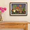 A framed art print depicting a whimsical vegetable harvesting scene, hanging on a beige wall above a wooden console table with a vase of pink flowers.