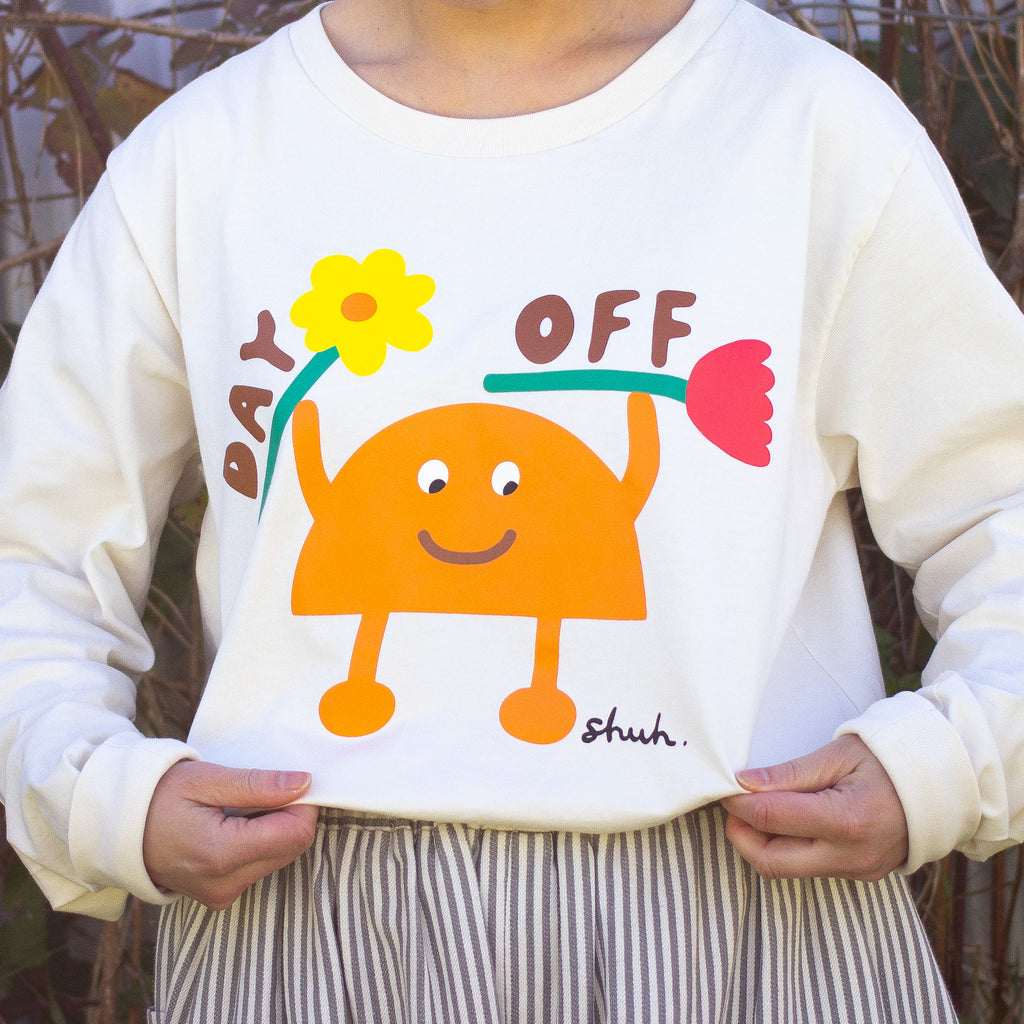Another front close up of a Person wearing a beige long sleeve t-shirt with a colourful graphic of an orange cartoon character holding two flowers and the words "DAY OFF" in playful text. Design by shuh., printed in Australia.