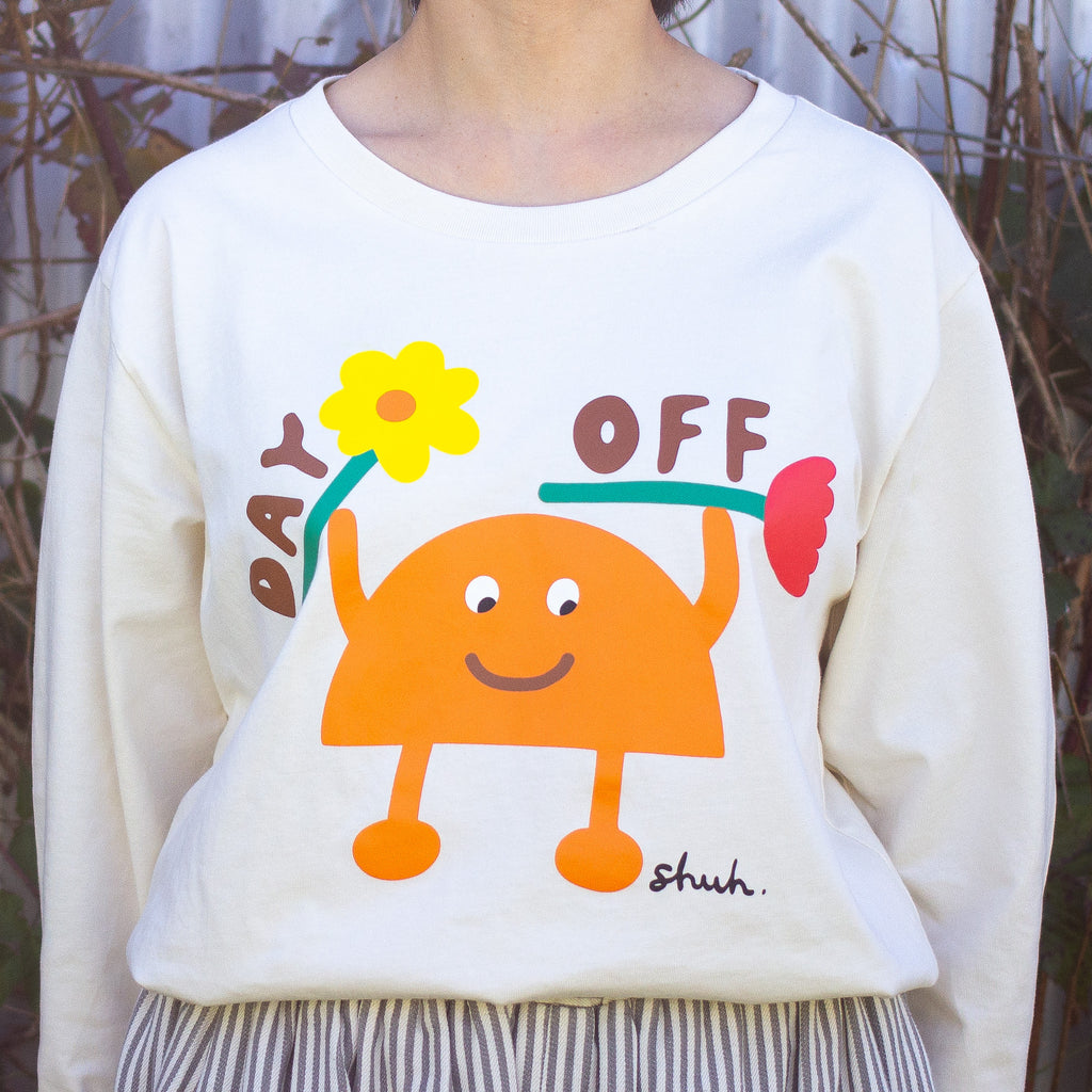 Front close up of Person wearing a beige long sleeve t-shirt with a colourful graphic of an orange cartoon character holding two flowers and the words "DAY OFF" in playful text. Design by shuh., printed in Australia.

