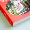 A close-up of a red paperback cover book lying on a light surface, with a circular printed photo of a forest path and red torii gate on the cover.