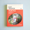 A book titled 'Day Tripper Tokyo' by Steve Wide and Michelle Mackintosh, featuring a coral red and cream cover with a circular photograph of a Japanese torii gate in a forest, set against a light blue background.