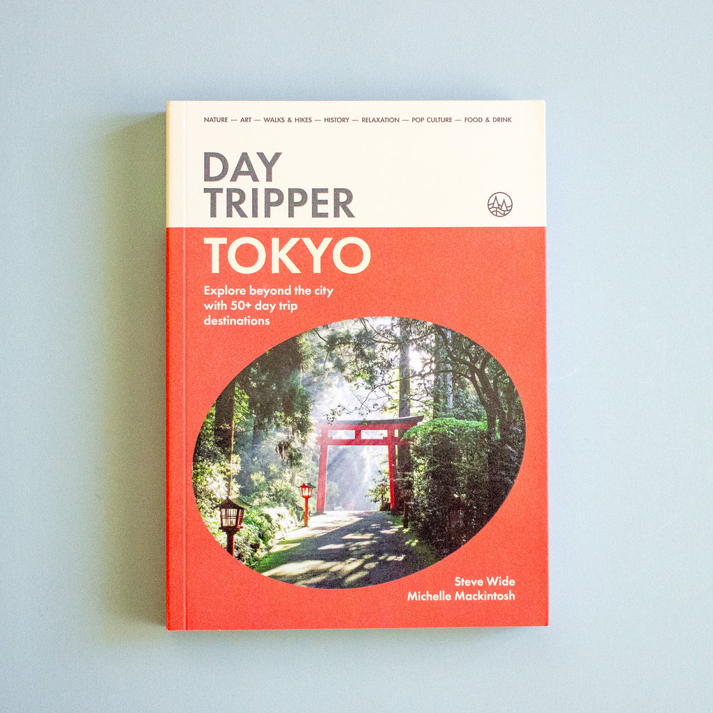 A book titled 'Day Tripper Tokyo' by Steve Wide and Michelle Mackintosh, featuring a coral red and cream cover with a circular photograph of a Japanese torii gate in a forest, set against a light blue background.