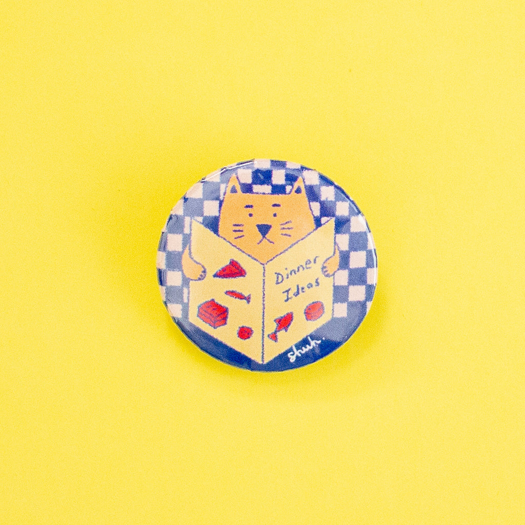 A round pin badge featuring a cartoon orange cat reading a book titled 'Dinner Ideas' with red food illustrations, set against a blue and white checkered background. The badge is displayed on a solid yellow background.